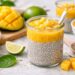 Chia Pudding Mango in Glas mit frischem Mangopüree, Chiasamen und geschnittenen Früchten auf heller Oberfläche angerichtet