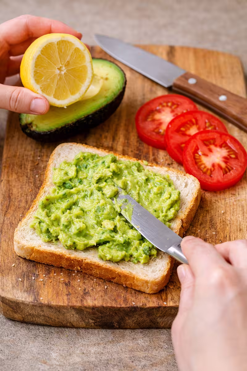 Avocado wird mit Zitronensaft auf Sandwichbrot gestrichen, daneben liegen Tomatenscheiben, Messer und Holzbrett
