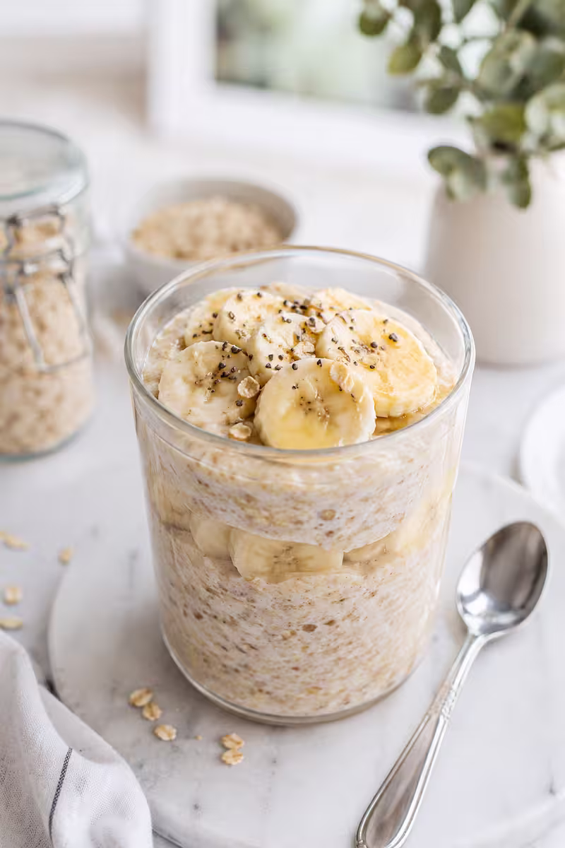 Cremige Overnight Oats Banane im Glas Cremige Overnight Oats Banane im Glas mit Bananenscheiben, Haferflocken und kleinem Löffel auf hellem Frühstückstisch