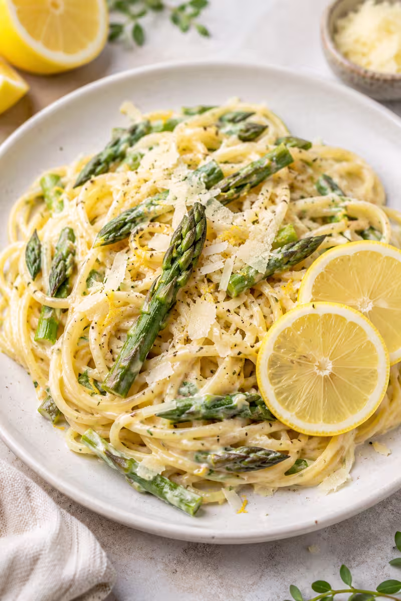 Cremige Zitronenpasta mit grünem Spargel auf hellem Teller mit Zitronenscheiben und Parmesan