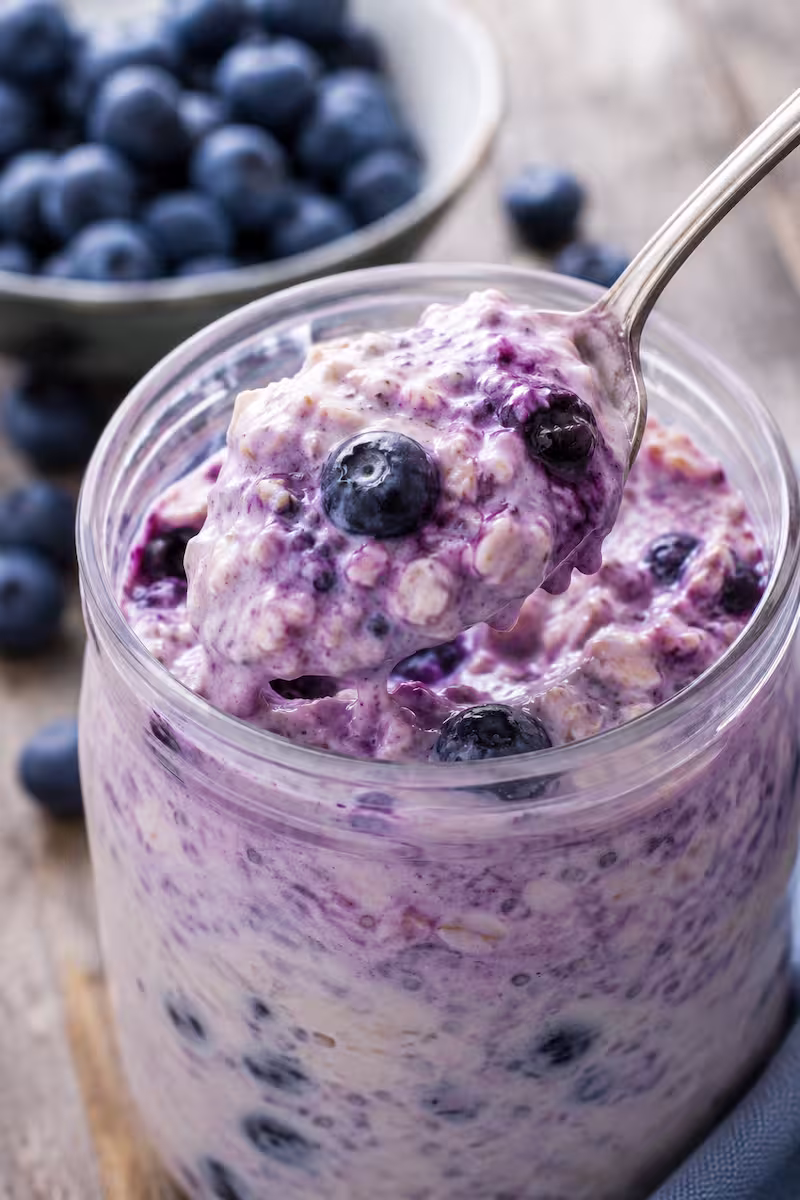 Löffel hebt cremige Blaubeer-Overnight-Oats aus einem Glas, Heidelbeeren im Hintergrund