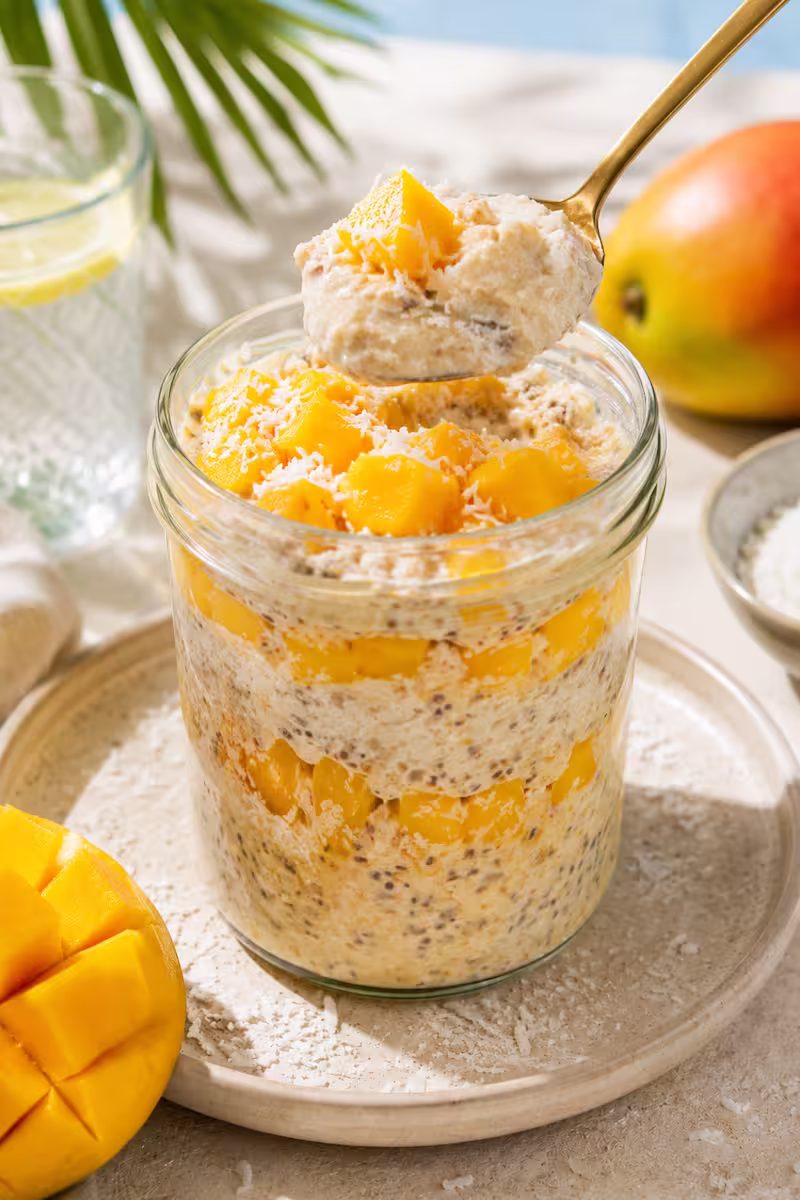 Löffel hebt cremige Mango Kokos Overnight Oats Löffel hebt cremige Mango Kokos Overnight Oats aus einem Glas, sichtbare Mangoschichten und Kokosraspel