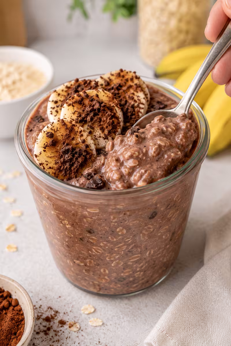 Löffel taucht in cremige Schoko-Bananen Overnight Oats mit sichtbaren Haferflocken und Schokosplittern