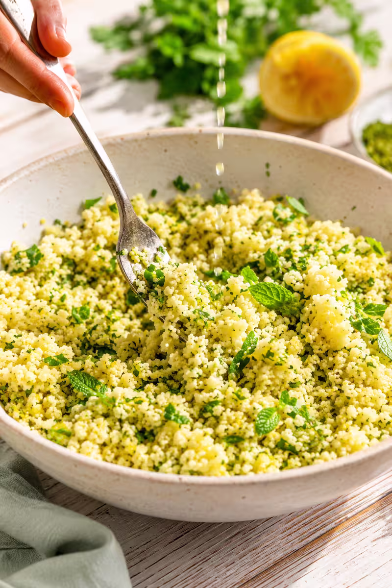 Nahaufnahme von lockerer Couscous-Struktur mit Gabelbewegung, frische Kräuter und Zitronensaft werden in Bowl eingerührt, sommerliche Food-Fotografie mit hellem Holzuntergrund