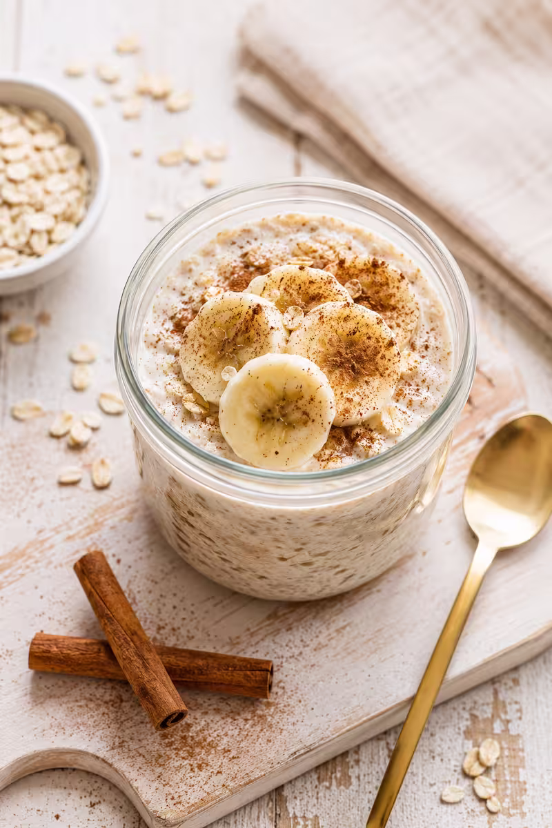 remige Overnight Oats mit Banane und Zimt im Glas, goldener Löffel, Haferflocken und Zimtstangen daneben