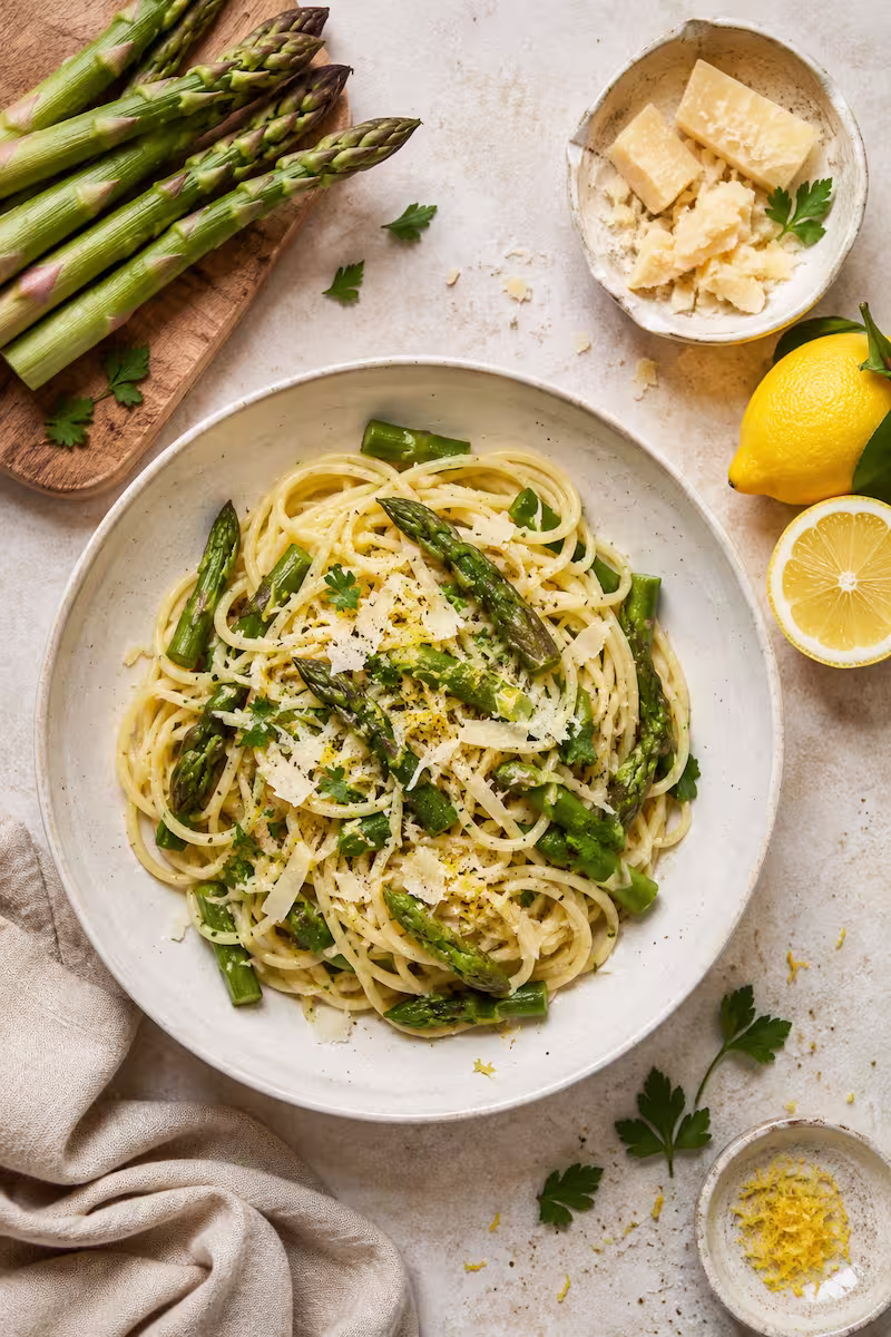 Top-Down-Aufnahme mit Pasta, grünem Spargel, Parmesan und frischer Zitrone auf hellem Küchentisch