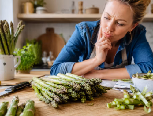 Frau in heller Küche betrachtet frischen grünen Spargel auf einem Holzbrett und vermeidet typische Fehler bei der Zubereitung – Grüner Spargel: Die häufigsten Fehler vermeiden