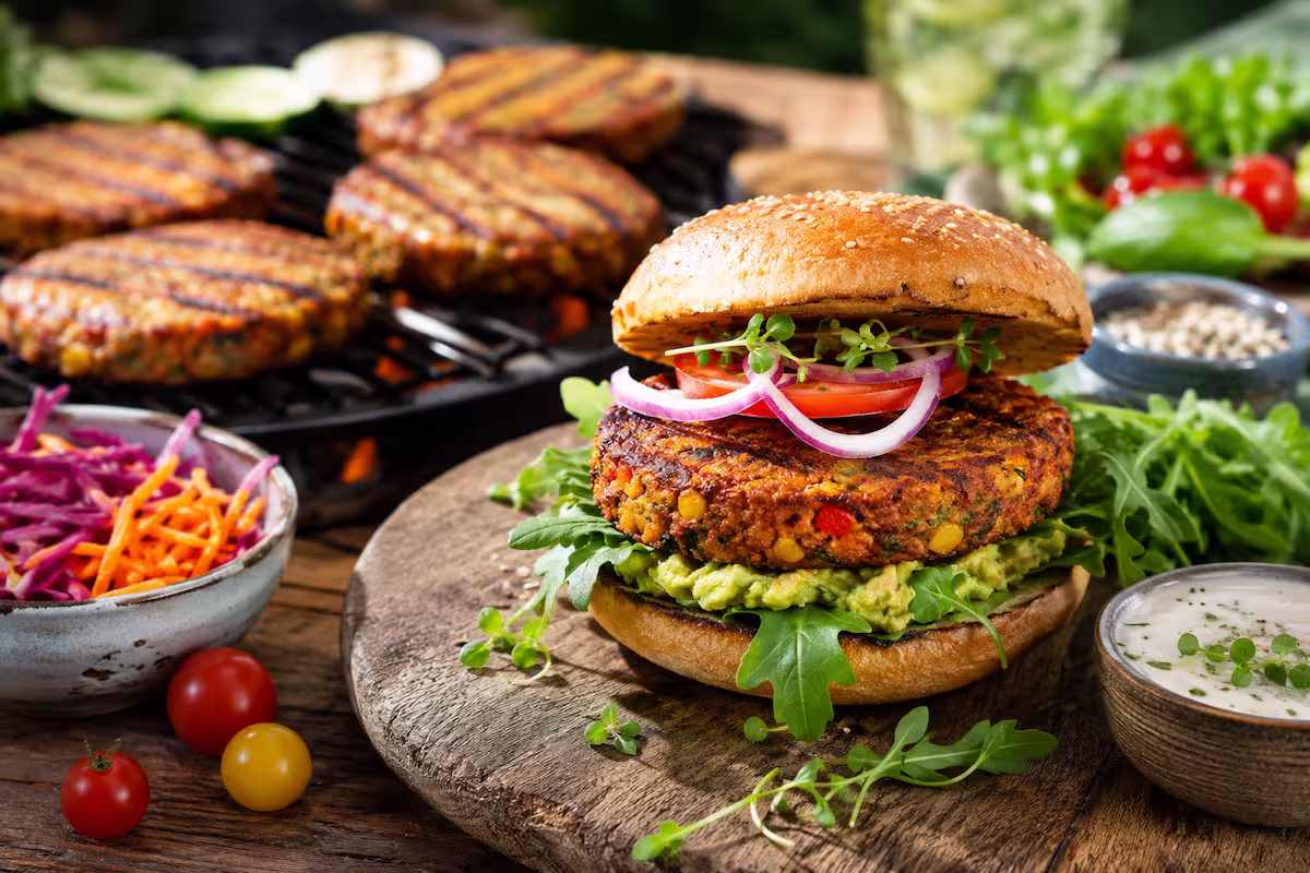 Gemüseburger Patties vom Grill mit knuspriger Kruste in einem Burgerbrötchen, belegt mit Salat, Tomaten und roten Zwiebeln, auf einem Holzteller vor einem Grill mit weiteren vegetarischen Burger-Patties und frischen Beilagen.